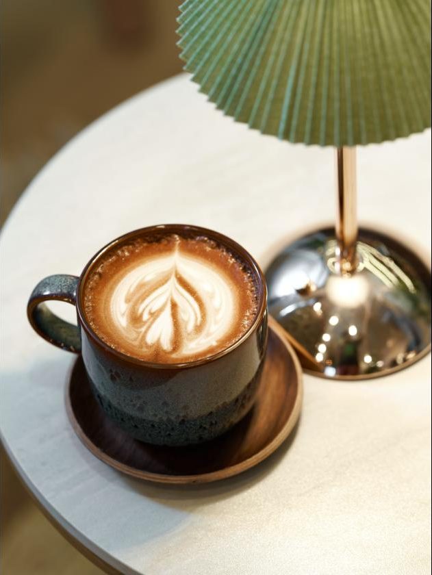 A latte in a dark glazed cup on a marble table, beside a brass and green lamp